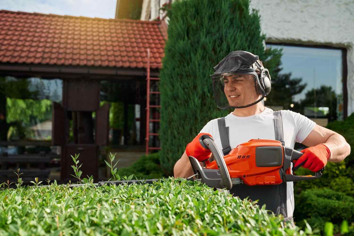 Landscaping professional in safety gear using a motorized trimmer to shape hedges.