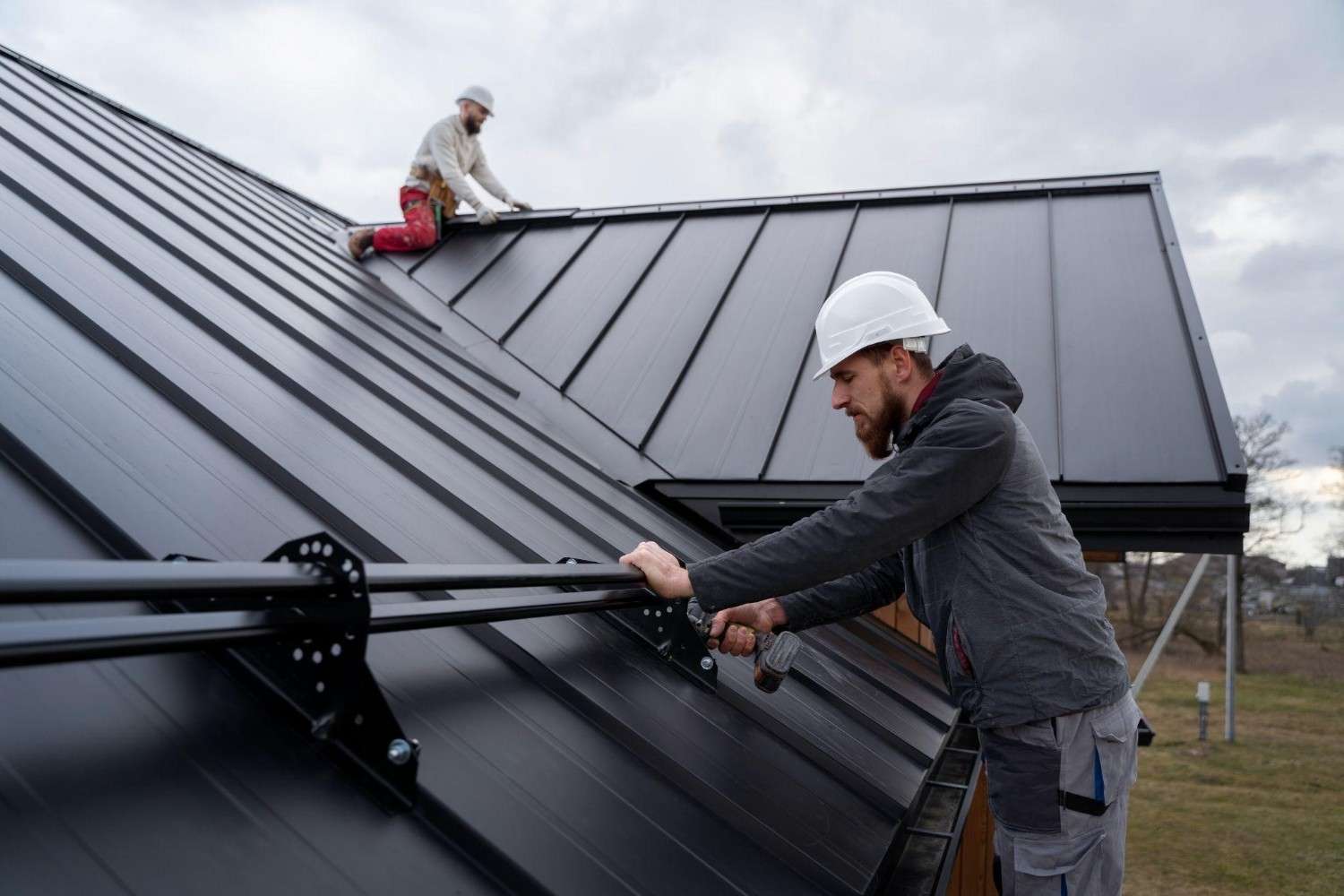 Two roofers in hard hats working on a black standing seam metal roof.
