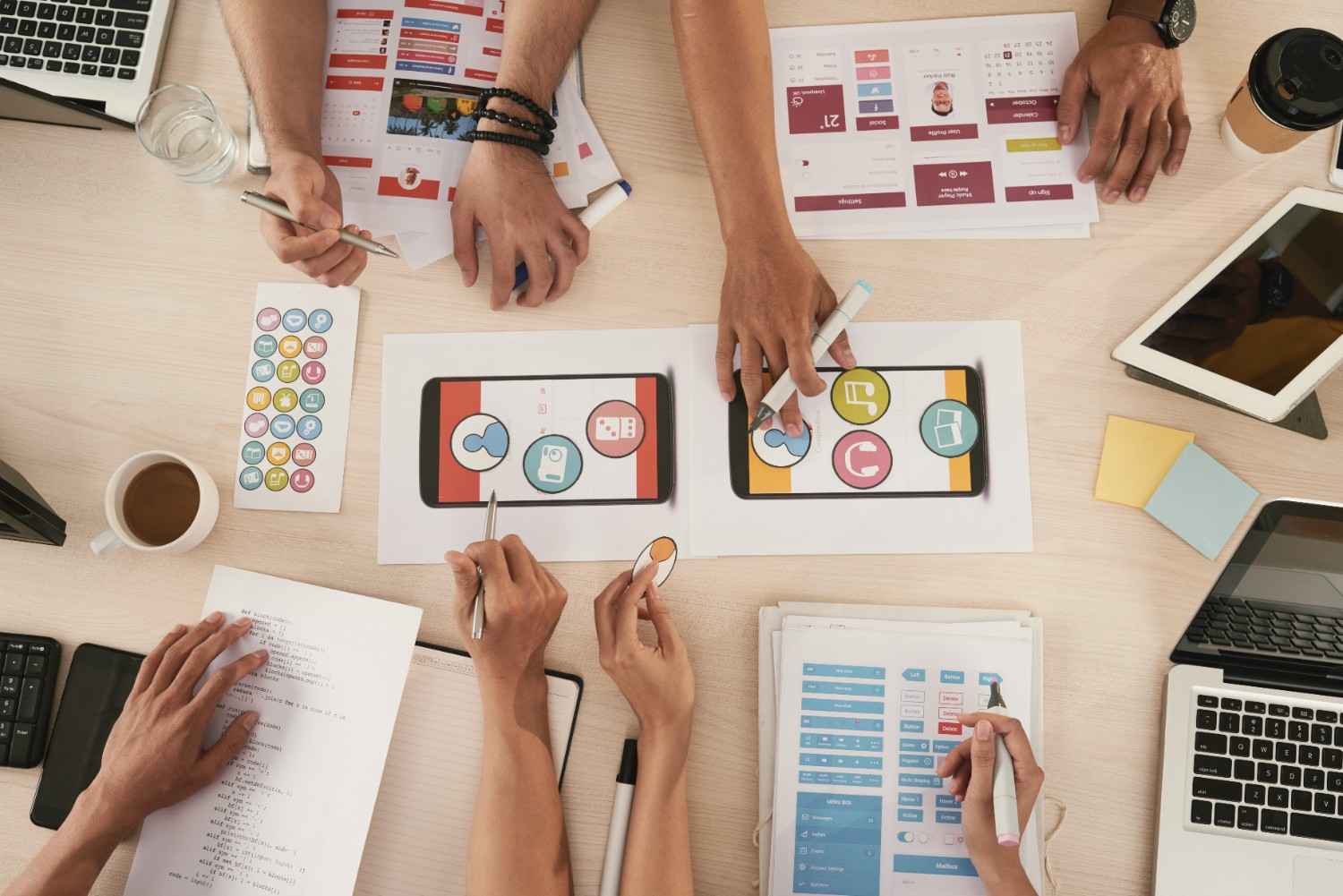 Top-down view of a digital agency team collaborating on mobile app UI/UX wireframes and web design layouts on a desk.