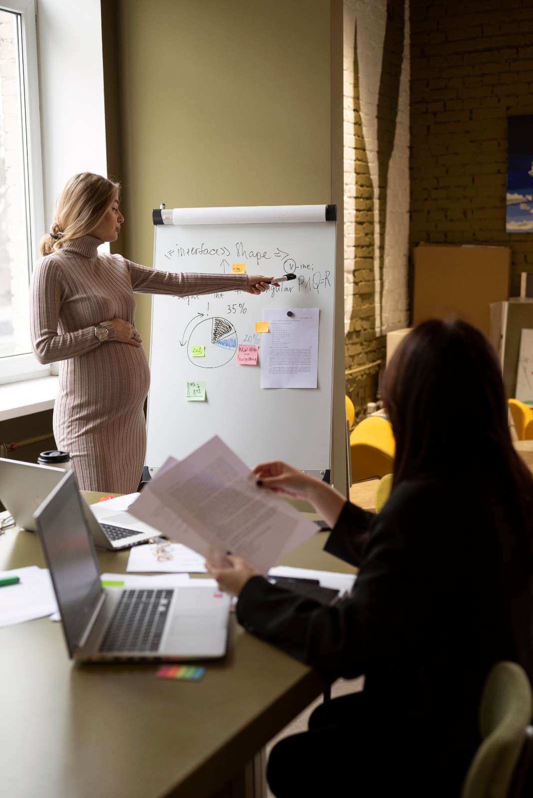 A professional woman presenting a data pie chart and strategy outline on a whiteboard during an office meeting.